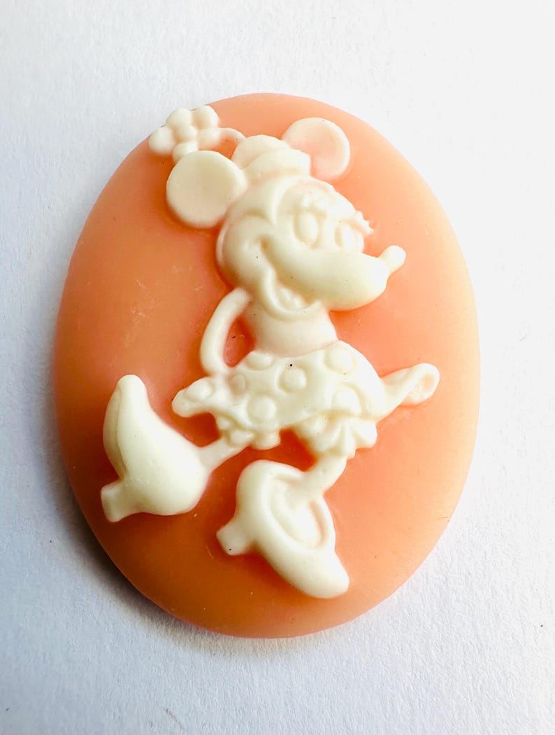 May include: A cameo featuring a cream-colored Minnie Mouse figure on a peach-colored oval background. Minnie Mouse is depicted in a classic pose, wearing a polka-dot skirt and a bow. The cameo is likely made of resin or a similar material.