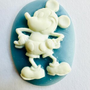 May include: A white Mickey Mouse figure is set against a light blue oval background. The cartoon character is in a standing pose with arms at his sides. The figure is a three-dimensional design.