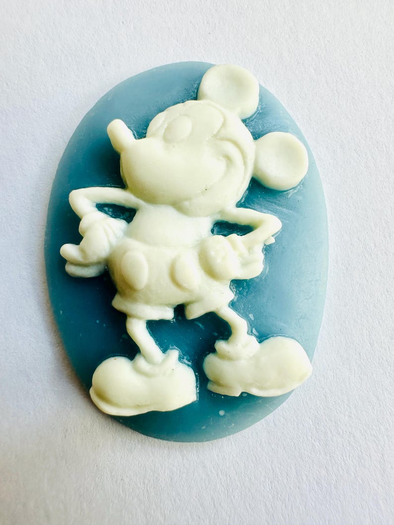 May include: A white Mickey Mouse figure is set against a light blue oval background. The cartoon character is in a standing pose with arms at his sides. The figure is a three-dimensional design.