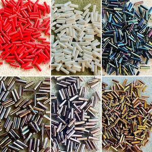 May include: A collection of six distinct piles of cylindrical bugle beads in various colors, including solid red, opaque white, iridescent dark blue, metallic silver, metallic purple, and metallic gold, each arranged in a separate grid section.