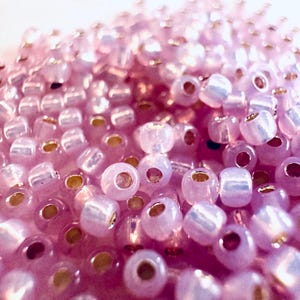 May include: Close-up of a pile of small, translucent, light purple beads. Each bead has a golden-colored hole. The beads are scattered, creating a textured surface. The image is well-lit, highlighting the details of the beads.