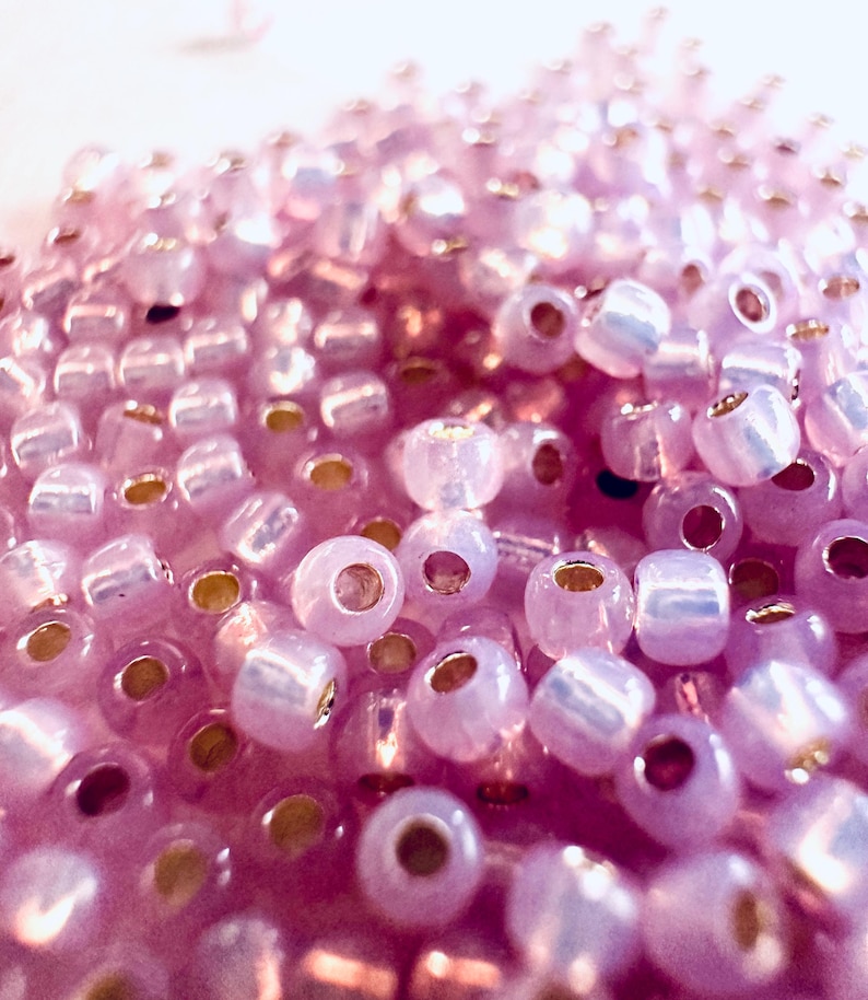 May include: Close-up of a pile of small, translucent, light purple beads. Each bead has a golden-colored hole. The beads are scattered, creating a textured surface. The image is well-lit, highlighting the details of the beads.