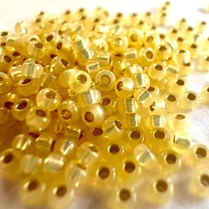 May include: A close-up shot of a pile of small, translucent yellow seed beads. The beads have a slightly iridescent quality, with a golden sheen. Each bead has a hole running through the center, and they are scattered across a white surface.