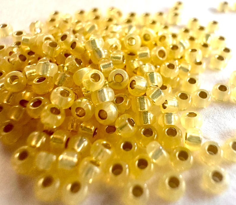 May include: A close-up shot of a pile of small, translucent yellow seed beads. The beads have a slightly iridescent quality, with a golden sheen. Each bead has a hole running through the center, and they are scattered across a white surface.