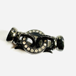 May include: A gunmetal-colored jewelry clasp with a decorative design. The clasp features two interlocking circular elements, each adorned with small, clear crystals. The clasp is designed to secure jewelry pieces, such as necklaces or bracelets.