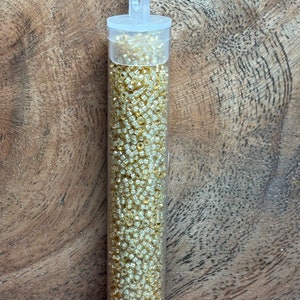 May include: A clear plastic tube filled with small, gold-colored seed beads. The tube has a hanging loop at the top.