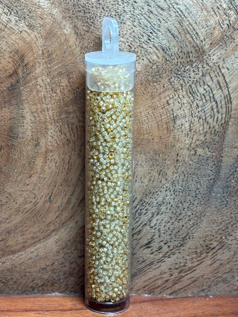 May include: A clear plastic tube filled with small, gold-colored seed beads. The tube has a hanging loop at the top.