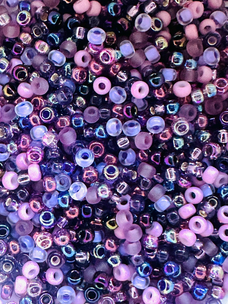 May include: A close-up of a large assortment of small, round beads in shades of purple, pink, and blue. The beads are made of glass and have a variety of finishes, including iridescent, matte, and clear.