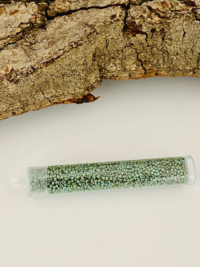 May include: A clear plastic tube filled with small, light green glass beads. The tube is lying on a white surface.