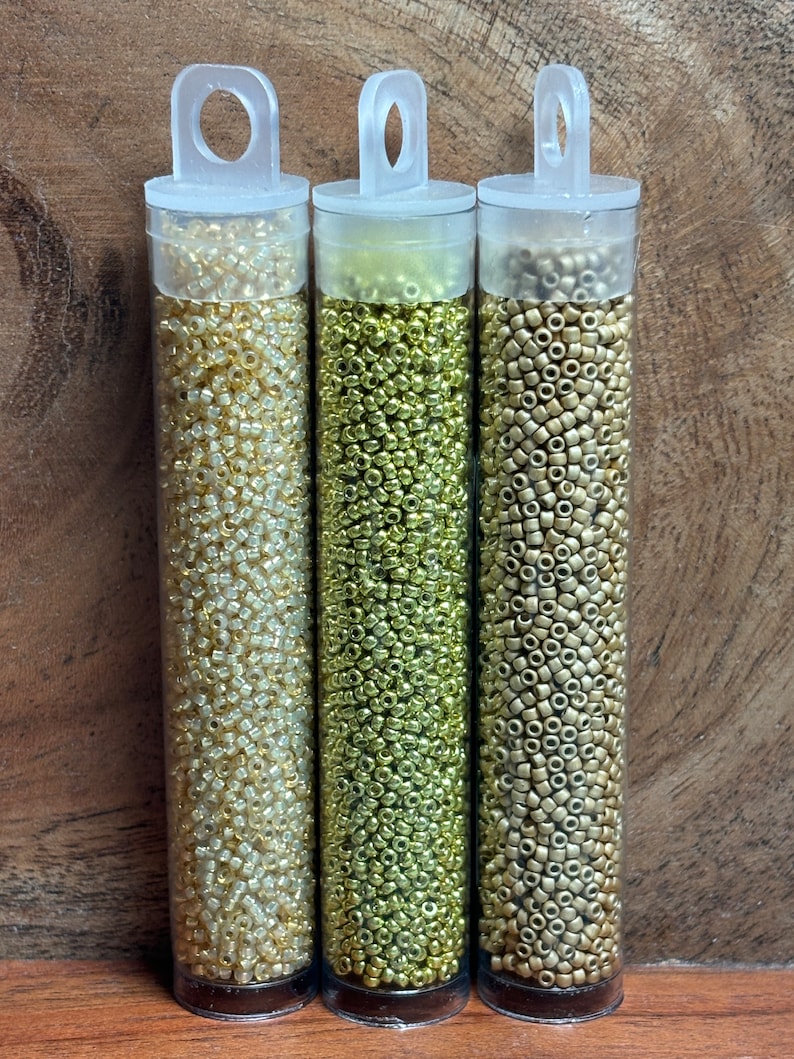 May include: Three clear plastic tubes filled with gold, silver, and bronze seed beads. The tubes have a clear plastic handle at the top.