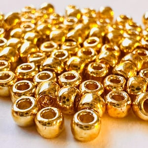 May include: Close-up of a pile of shiny, golden-colored beads. The beads are cylindrical with a hole running through the center, reflecting light. These beads are suitable for jewelry making and crafting.