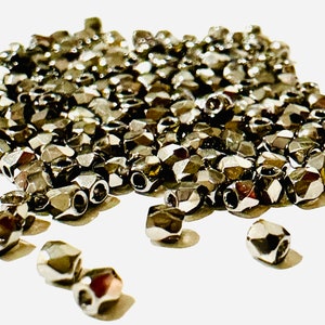 May include: A pile of silver-colored metal beads with a faceted, geometric shape. The beads are small and have a hole in the center for stringing.
