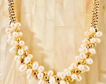May include: A gold-toned necklace featuring a floral design. The necklace is composed of white pearls and gold-colored beads. The chain is a woven gold color. The necklace is displayed against a cream-colored background.