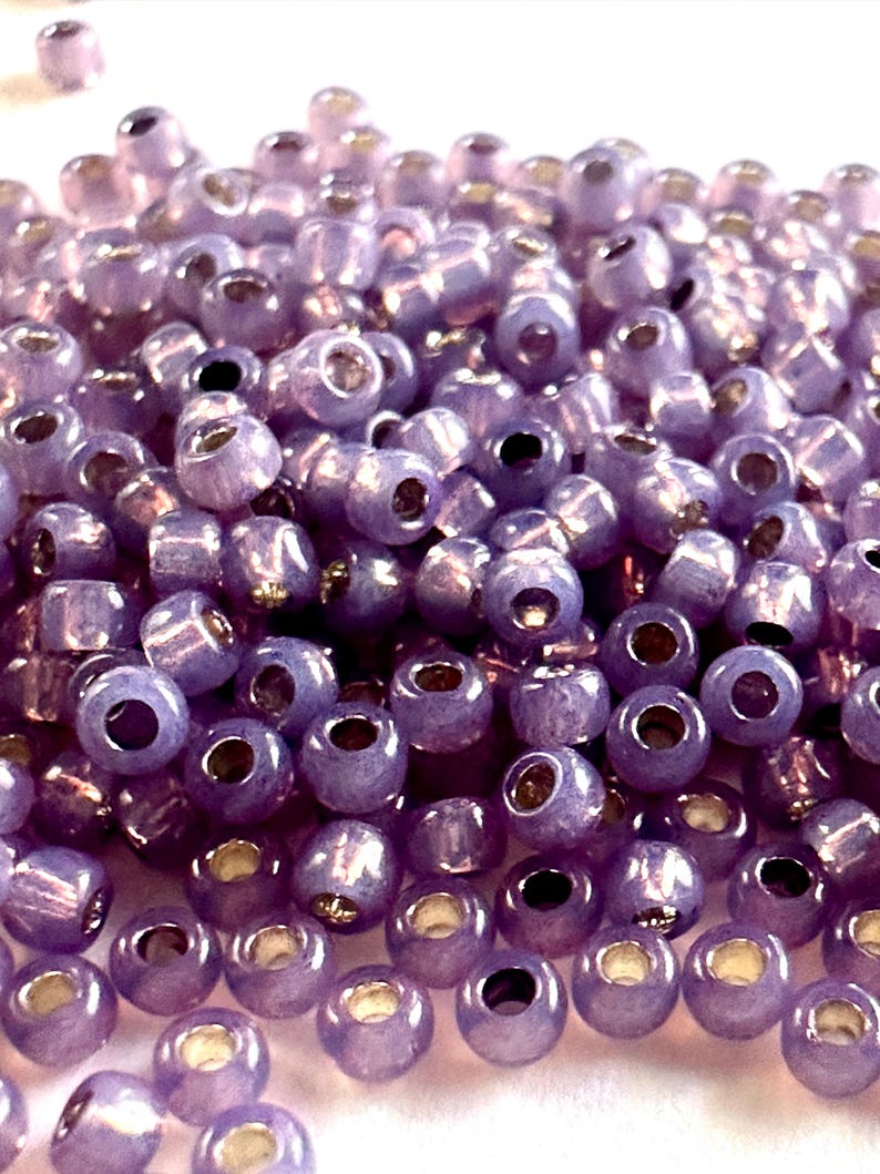 May include: Close-up of a pile of small, round, lavender-colored beads. Each bead has a hole running through the center and a metallic gold-colored interior. The beads are scattered, creating a textured surface.
