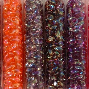 May include: Five tubes of iridescent glass beads. The tubes are filled with beads in shades of purple, red, and orange.