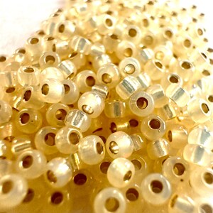 May include: Close-up of a pile of small, translucent, off-white beads with gold-colored cylindrical centers. The beads are scattered, creating a textured surface. Ideal for jewelry making and crafts.