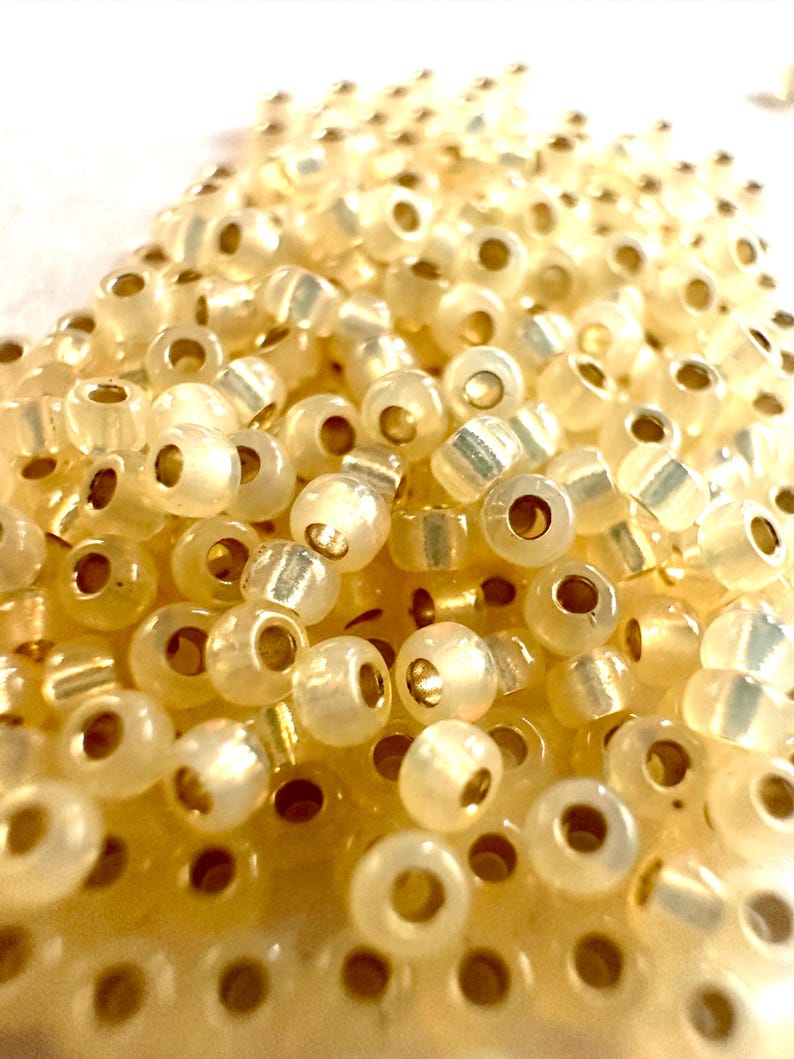 May include: Close-up of a pile of small, translucent, off-white beads with gold-colored cylindrical centers. The beads are scattered, creating a textured surface. Ideal for jewelry making and crafts.