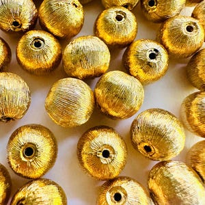 May include: A collection of gold-colored round beads with a textured surface. The beads are scattered on a white background.
