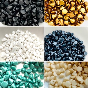 May include: A collection of six piles of small, triangular-shaped beads in various colors including matte black, metallic gold, solid white, dark navy blue, teal, and cream. Each bead features a flat circular top and a single hole for threading.