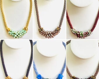 May include: Six beaded necklaces displayed on white busts. The necklaces feature a variety of colors, including gold, brown, red, black, and blue. Each necklace has a central cluster of beads in complementary colors, creating a unique design.