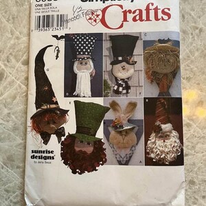 May include: A Simplicity Crafts sewing pattern envelope, number 8935, featuring illustrations of various fabric doll heads with hats. Designs include a witch, a patriotic figure, a snowman, a scarecrow, a leprechaun, a rabbit, and a bearded figure.