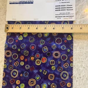May include: A sample of fabric with a dark blue background and a pattern of colorful buttons. A wooden ruler is placed across the fabric, and a white label with text is visible. The fabric is labeled "Horizons II".