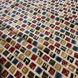 May include: A beige fabric with a repeating pattern of squares containing letters in black, red, blue, and brown. The letters are in a variety of fonts and sizes.