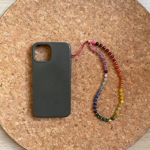 Può includere: Una custodia per telefono grigio scuro con un cinturino per telefono con perline arcobaleno. Il cinturino presenta una varietà di perline colorate, tra cui rosa, viola, blu, verde, giallo e arancione. La custodia e il cinturino del telefono sono su una superficie rotonda di sughero.