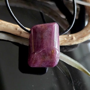 Könnte beinhalten: Ein polierter, rechteckiger Rubinanhänger mit dunkelroter Farbe, der an einer schwarzen Lederschnur befestigt ist. Der Edelstein weist natürliche Farb- und Texturvariationen auf. Die Halskette ist ein Schmuckstück.