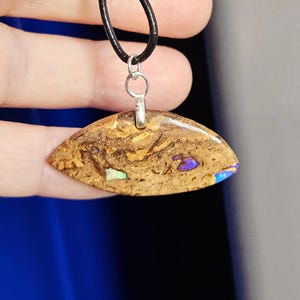 Puede incluir: Un collar con colgante de piedra pulida en forma de lágrima, con un patrón marrón y beige. La piedra tiene acentos iridiscentes azules y verdes. El colgante está sujeto a un cordón negro con una fianza plateada.