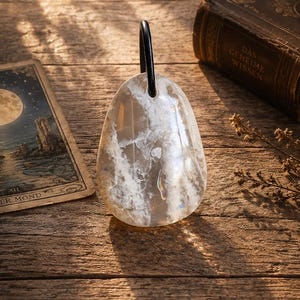 May include: A teardrop-shaped pendant made of clear, polished stone with white, cloud-like inclusions throughout the surface. The pendant features a smooth, rounded base and is suspended by a simple, thin black cord loop at the top center.