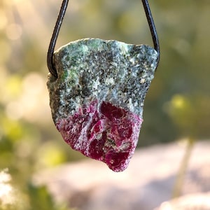 Op de afbeelding: Een ruwe, ongepolijste hanger met een diep magenta robijnkristal ingebed in een textuurrijke, gevlekte groene en grijze zoisietmatrix, hangend aan een eenvoudig zwart leren koord dat door een geboord gat boven in de steen loopt.