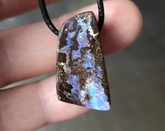 May include: A pendant necklace featuring a rectangular, polished gemstone with a black leather cord. The gemstone displays a mix of brown, blue, and white colors. The stone has a unique, natural pattern.