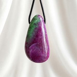 Può includere: Un ciondolo a forma di goccia realizzato in pietra di rubino zoisite lucida con motivi di colore magenta vibrante e verde intenso. La gemma liscia e lucida è sospesa a un semplice cordino nero, mostrando una texture naturale della pietra.