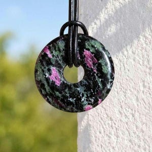 Könnte beinhalten: Ein runder Anhänger aus poliertem Rubin-Zoisit-Stein mit einem gesprenkelten Muster aus dunkelgrünen, schwarzen und leuchtend magentafarbenen Einschlüssen. Der Stein ist wie ein flacher Donut geformt und an einem schwarzen Lederband befestigt.