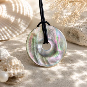 Puede incluir: Un colgante circular hecho de nácar iridiscente con un orificio central, suspendido de un cordón de cuero negro. La superficie muestra tonos arremolinados de verde, rosa y plata, reflejando la luz con un brillo nacarado y multicolor.