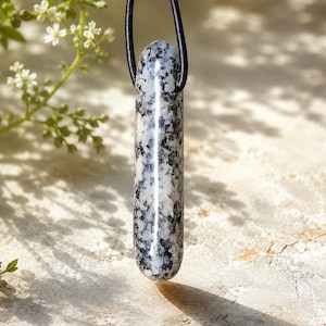 Pode incluir: Um pingente cilíndrico com um cordão de couro preto. O pingente é feito de uma pedra salpicada branca e preta. A pedra tem uma superfície polida e lisa. O pingente está pendurado em frente a um fundo neutro.