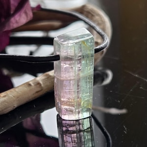 May include: A rectangular, multi-colored tourmaline gemstone pendant. The gemstone is a mix of pink, green, and clear hues, suspended on a black cord. The pendant is displayed against a dark, reflective surface, with a blurred background.