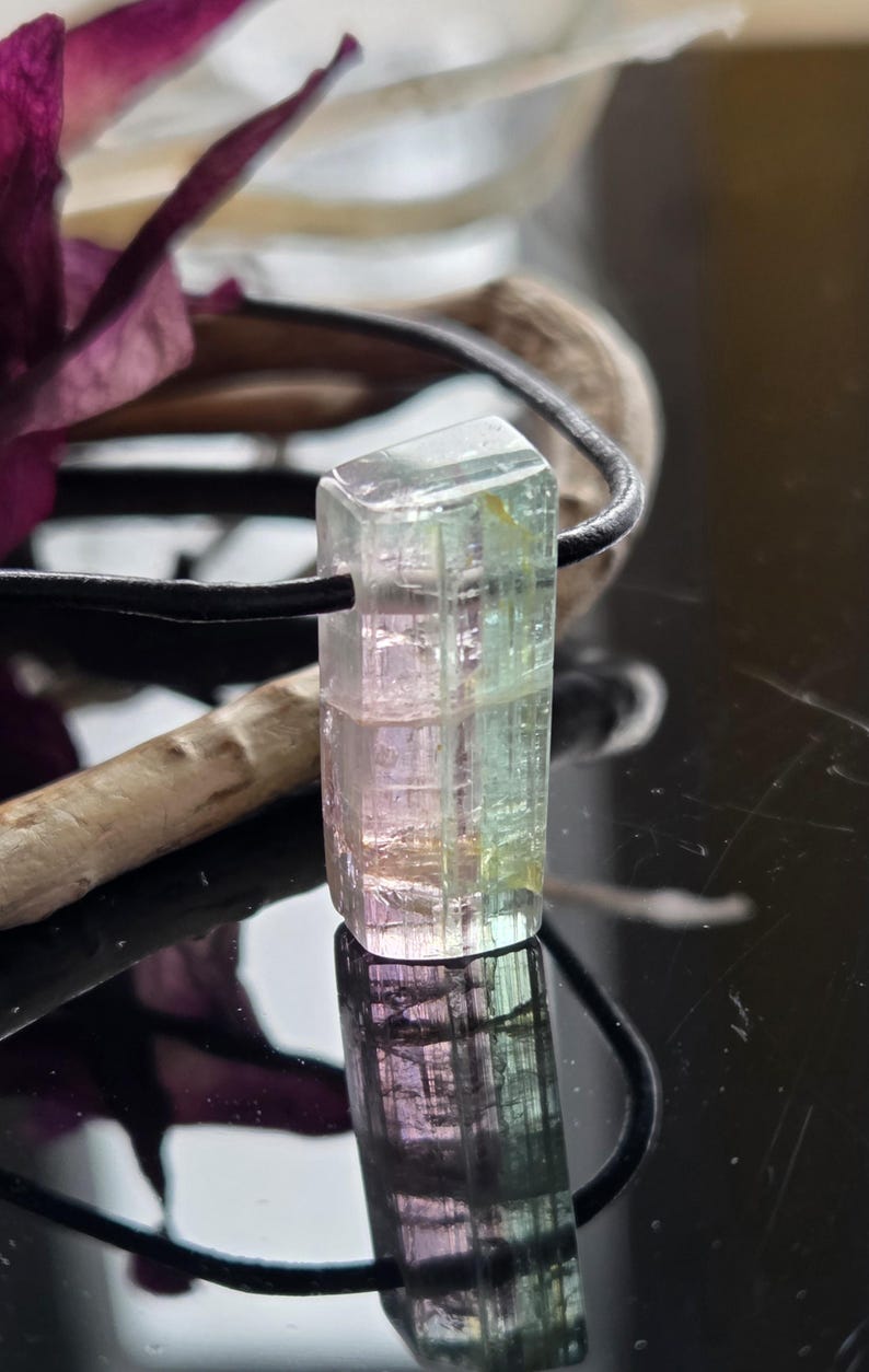 May include: A rectangular, multi-colored tourmaline gemstone pendant. The gemstone is a mix of pink, green, and clear hues, suspended on a black cord. The pendant is displayed against a dark, reflective surface, with a blurred background.