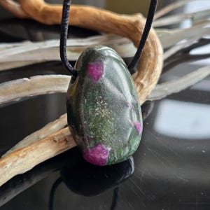 Könnte beinhalten: Halskette mit einem ovalen Anhänger aus grünem Stein mit rosa Einschlüssen. Der Anhänger hängt an einer schwarzen Kordel. Der Hintergrund zeigt ein Stück Treibholz. Der Stein ist etwa 3 cm lang.