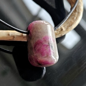 May include: A pink and white gemstone pendant on a black leather cord. The pendant is oval shaped and has a smooth, polished finish.