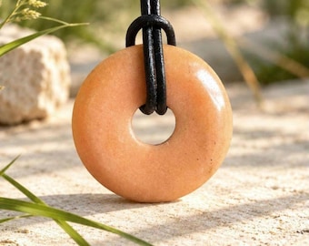 May include: A smooth, circular peach-colored stone pendant with a central hole, suspended from a black leather cord. The pendant features a polished, uniform surface and a rounded, donut-like shape, hanging vertically from the dark cord.