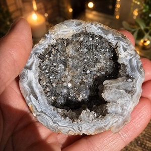 Könnte beinhalten: Eine kreisförmige Achat-Geode mit einem hohlen Zentrum, das mit kleinen, funkelnden klaren und grauen Quarzkristallen gefüllt ist. Der äußere Rand zeigt konzentrische Bänder aus weißen, grauen und beigen Mineralschichten, die eine natürliche Steinform bilden.