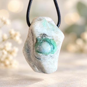 Può includere: Un ciondolo composto da una pietra grezza di forma irregolare, bianca con evidenti inclusioni minerali verdi e un centro liscio e arrotondato. La pietra è sospesa a un semplice cordino nero, mostrando una consistenza naturale e grezza.