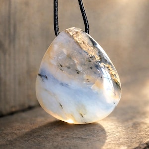 Può includere: Una collana con pendente con una pietra lucida a forma di goccia. La pietra mostra un mix di colori, tra cui bianco, blu, beige e nero, con una finitura lucida. Il pendente è sospeso a un cordino nero.