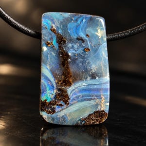 Peut inclure: Un pendentif rectangulaire composé d'une pierre d'opale bleue polie aux motifs tourbillonnants bleu clair et turquoise, parsemée d'inclusions minérales brun foncé et bronze métallique, suspendu à un simple collier en cordon noir.