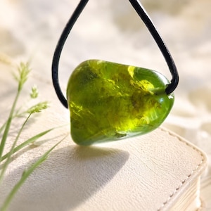 Puede incluir: Un colgante de piedra preciosa verde oliva translúcida, suspendido de un cordón negro. La piedra pulida tiene una forma redondeada e irregular. El colgante se encuentra sobre un fondo suave y neutro, con un toque de vegetación en primer plano.