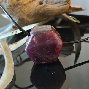 Könnte beinhalten: Ein facettierter, tiefvioletter Edelsteinanhänger an einer schwarzen Kordel. Der Edelstein hat eine transparente Qualität mit sichtbaren inneren Mustern. Der Anhänger wird auf einer reflektierenden Oberfläche mit Treibholz im Hintergrund präsentiert.