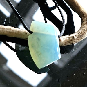Può includere: Una collana con pendente azzurro e verde e cordino nero. Il pendente è di forma rettangolare con bordi arrotondati. La collana è esposta su un pezzo di legno trasportato dalle correnti.