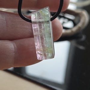 May include: A rectangular pendant necklace featuring a multi-colored tourmaline gemstone. The gemstone displays pink, green, and clear sections. It is suspended from a black cord. The pendant is approximately 1 inch long.